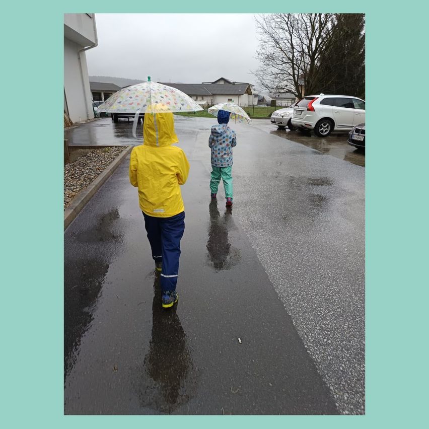 Zwei Kinder gehen auf einer nassen Straße bei Regen und halten Regenschirme. Sie tragen Regenmäntel und Turnschuhe. Hinter ihnen sind geparkte Autos und ein Haus.
