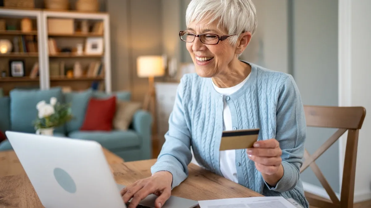 An elderly woman smiles while using a laptop and holding a credit card, likely shopping online. She is wearing glasses and a light blue sweater.