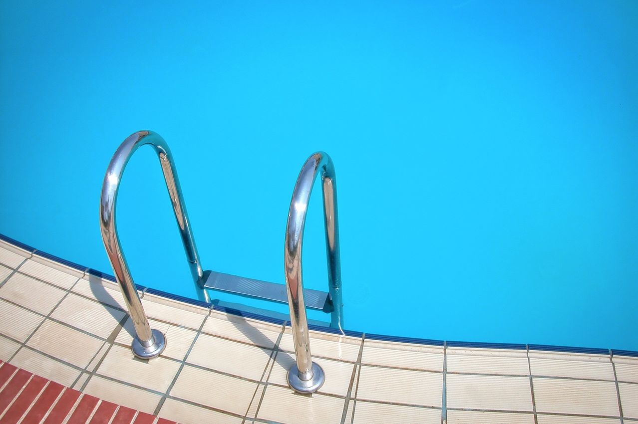 Ein Handlauf aus Edelstahl verläuft entlang der Seite eines blauen Schwimmbeckens. Die Poolfliesen haben einen hellbeigen und roten Rand.
