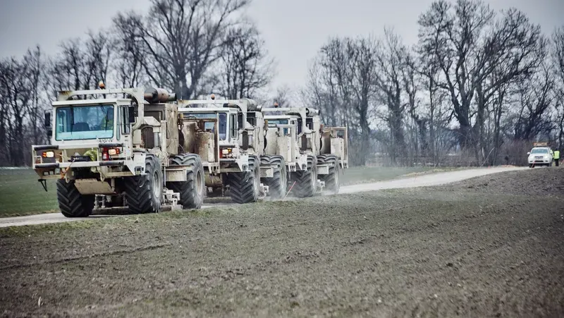 Several large trucks drive down a dirt road, with a car following behind. The area is surrounded by barren trees.