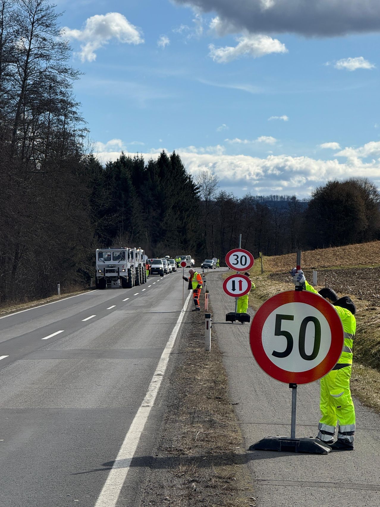Eine Straßenbaumannschaft installiert ein Geschwindigkeitsschild, während Arbeiter in Warnwesten daneben stehen. Fahrzeuge sind am Straßenrand geparkt, und Bäume säumen die Straße. Der Himmel ist teilweise bewölkt.