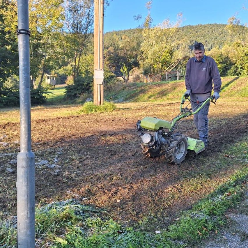 Ein Mann benutzt einen grünen Traktor auf einem Grasfeld mit einem Metallpfosten in der Nähe. Die Umgebung hat Bäume und einen Zaun.