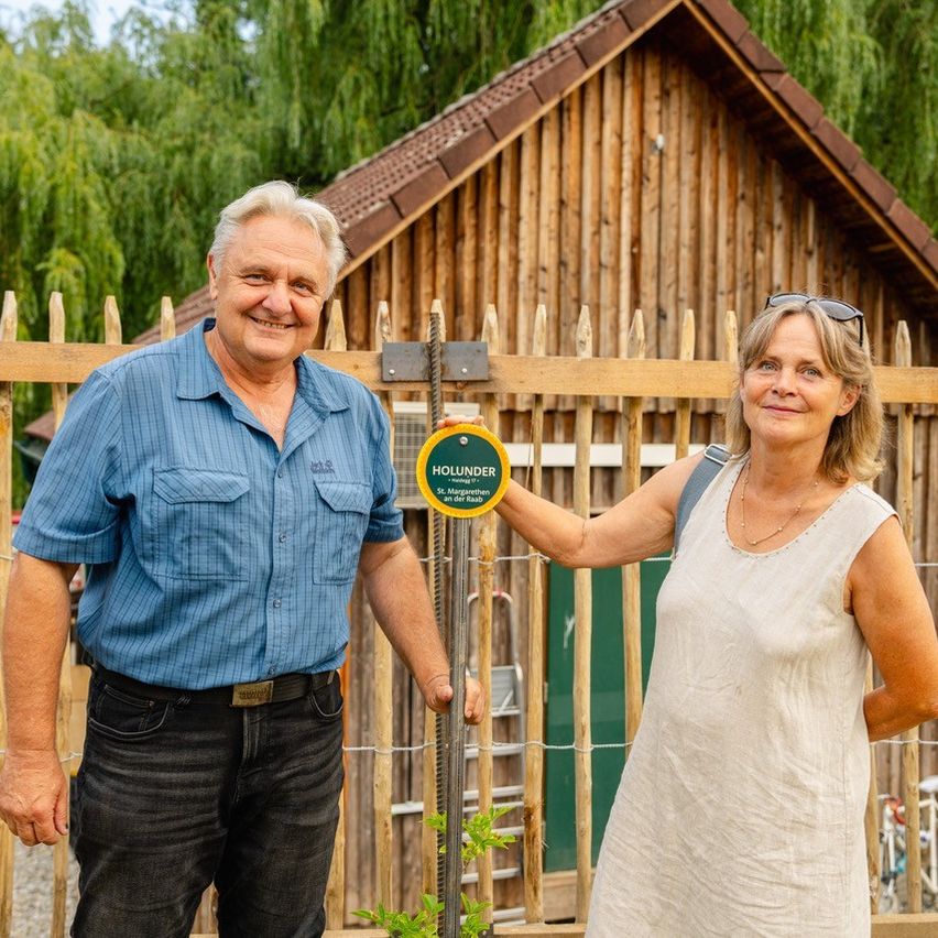 Ein älterer Mann und eine Frau stehen neben einem Holzzaun und lächeln. Der Mann trägt ein blaues Hemd und schwarze Hose, während die Frau ein weißes Kleid trägt und einen Rucksack hat. Ein Schild mit dem Wort 'HOUNDER' ist am Zaun befestigt.
