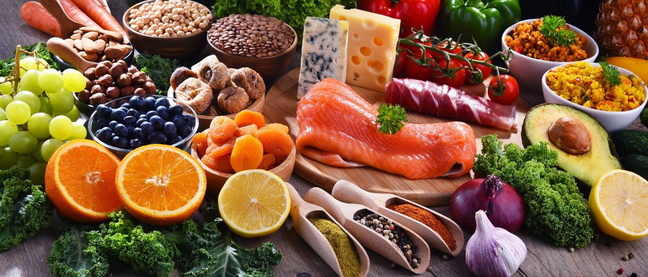 Auf einem Holztisch sind verschiedene Lebensmittel ausgestellt: ein Lachsfilet, Käse, Tomaten, Paprika, Zwiebeln, Knoblauch, Blaubeeren, getrocknete Aprikosen, Pilze und Hülsenfrüchte, zusammen mit einer halben Zitrone und Orange. Gewürze und Kräuter sind in hölzernen Schöpfchen.