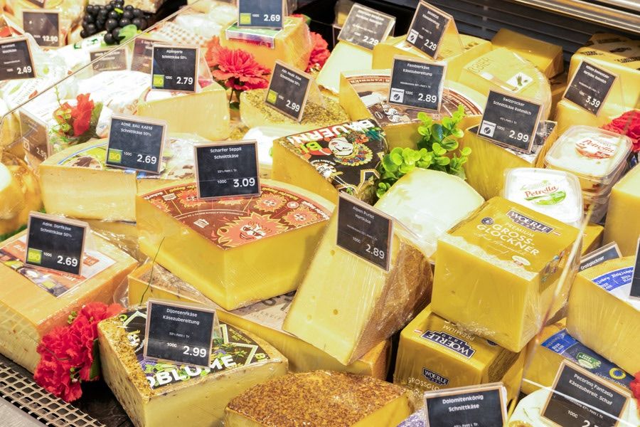 A cheese shop display with various cheeses in plastic packaging. Each cheese has a price tag and some have flowers. The cheeses are arranged in layers.