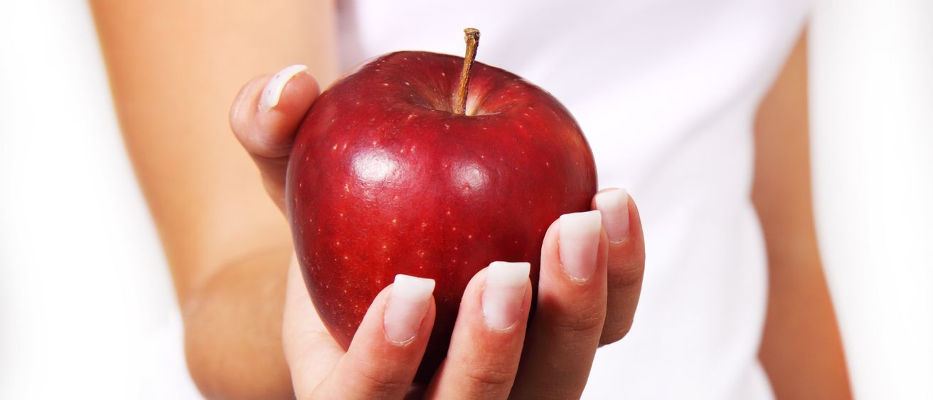 Eine Person hält einen roten Apfel vor einem weißen Hintergrund. Die Nägel der Person sind weiß lackiert.