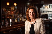 Eine lächelnde Frau mit roten Haaren sitzt an einer Bartheke, mit einer Reihe von Alkoholflaschen hinter ihr und einem unscharfen Hintergrund eines Bar-Interieurs.