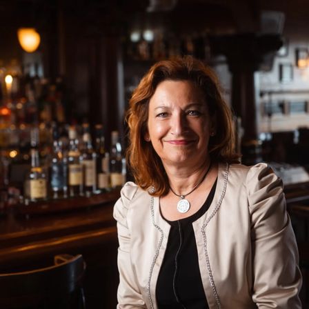 Eine Frau mit roten Haaren lächelt in einer Bar, mit Regalen voller Likörflaschen im Hintergrund.
