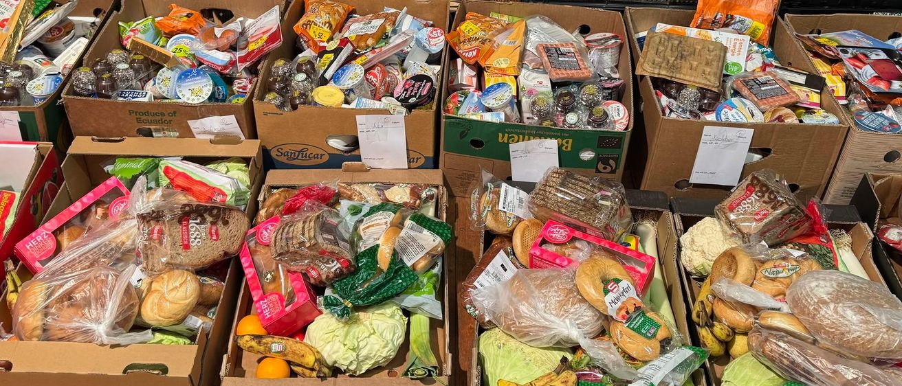 Ein Holzregal mit mehreren Kartons, die mit verschiedenen Lebensmitteln wie Brot, Obst und Gemüse gefüllt sind. Die Kartons sind ordentlich in Reihen angeordnet, wobei einige Artikel in Plastik eingewickelt sind.