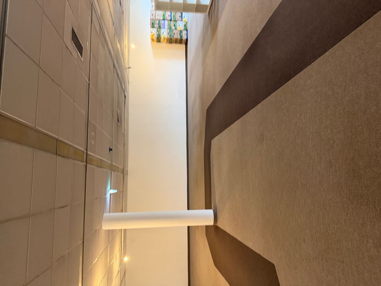 A hallway with tiled walls and a white pipe on the floor, leading to a room with a shelf and colorful boxes.