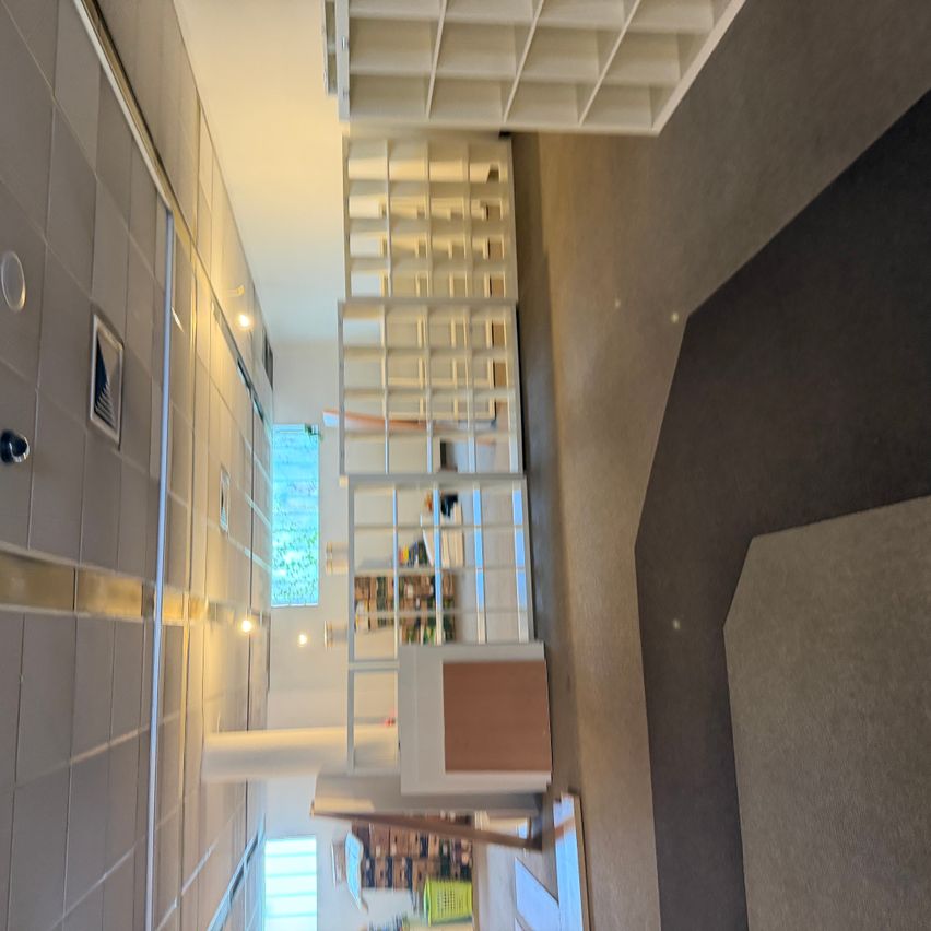 An aerial view of a library with white shelves, a tiled wall, and a carpeted floor. The shelves are filled with books, and there is a window with a view of trees outside.