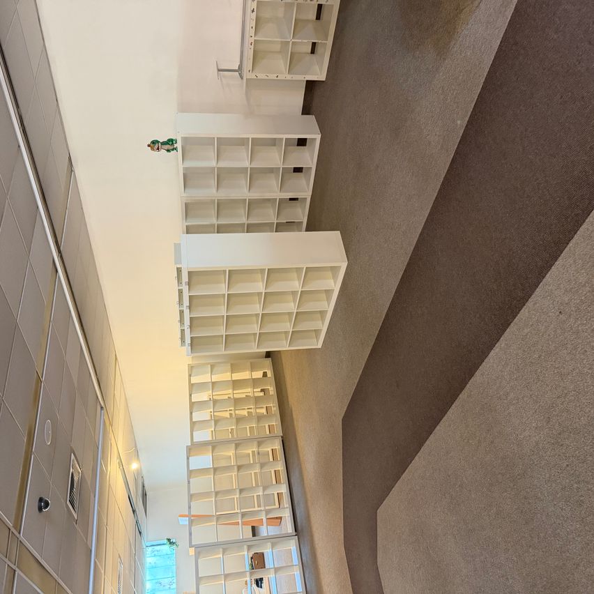 An aerial view of a room with white shelves and a brown carpeted floor. The shelves are neatly arranged in a row, and a green toy is placed on the top shelf. The room has a white wall and a gray ceiling.