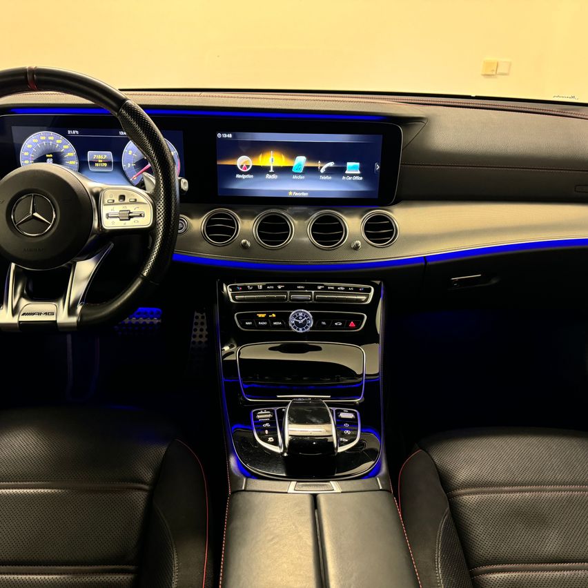 Innenraum eines Mercedes-Benz Autos mit einem großen Touchscreen-Display, schwarzen Ledersitzen, einem Lenkrad und blauer LED-Beleuchtung.