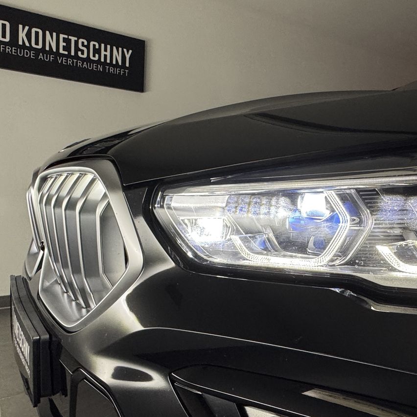 Nahaufnahme der Vorderseite eines schwarzen Autos, das Scheinwerfer und Kühlergrill zeigt. Der Stoßfänger des Autos hat ein Nummernschild. Hinter dem Auto befindet sich ein Schild mit der Aufschrift 'O KONETSCHNY FREUDE AUF VERTAURE TRIFFT.'