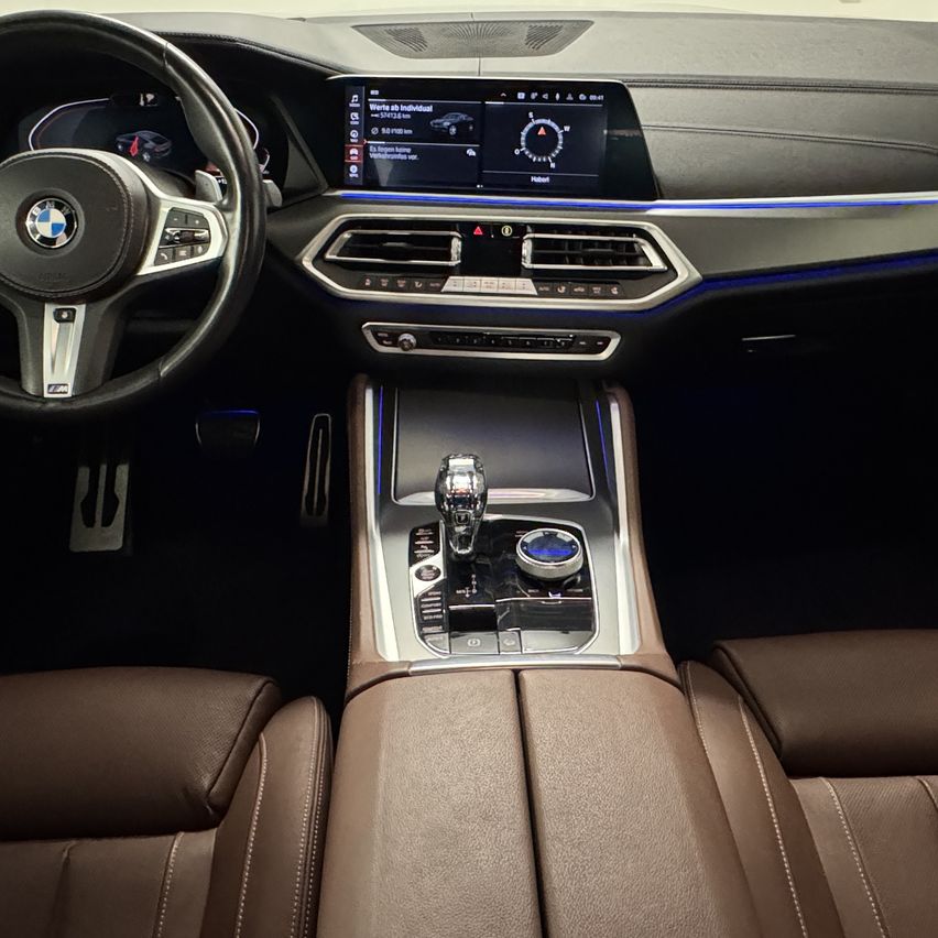 Innenraum eines BMW X7 SUV. Das Armaturenbrett zeigt einen Tachometer und verschiedene Fahrzeuginformationen an. Die Mittelkonsole verfügt über einen Gangschalter, Lüftungsöffnungen und ein Bedienfeld. Die Sitze sind aus braunem Leder.