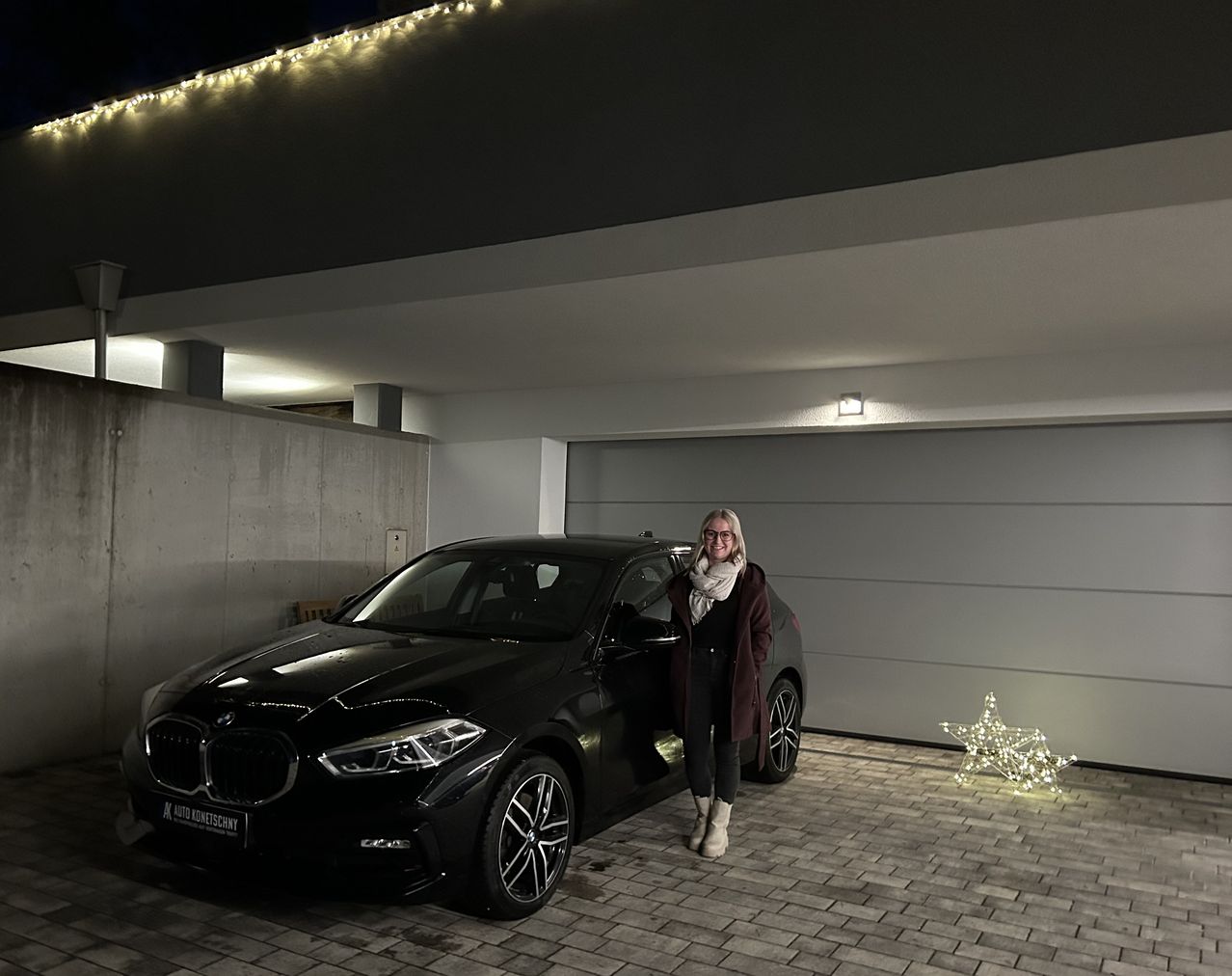 Eine Frau steht neben einem schwarzen BMW-Auto in einer Garage, beleuchtet von sternförmigen Lichtern. Das Auto hat ein Nummernschild, das 'K AUTO GOMETSCHNY' liest.