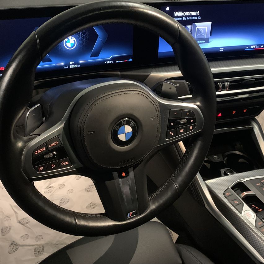 Das Innere eines BMW-Autos mit einem Lenkrad mit Steuertasten und einem Armaturenbrett-Bildschirm, der 'Willkommen!' und andere Informationen anzeigt.