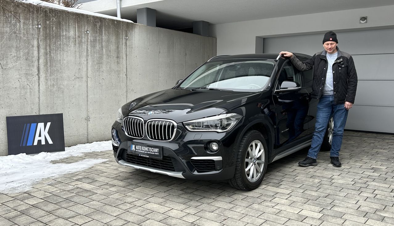 Ein Mann steht neben einem schwarzen BMW X1 in einer Garage, möglicherweise um einzusteigen. Das Fahrzeug hat ein Nummernschild mit der Aufschrift AK AUTO KONETSCHNY.
