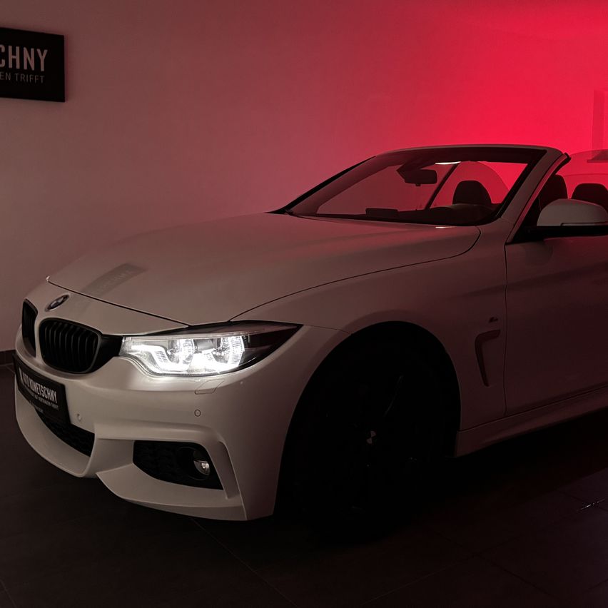 A white BMW 4 Series convertible is parked in a showroom with a red light illuminating the background. The car has a sleek design and is facing forward with headlights on.