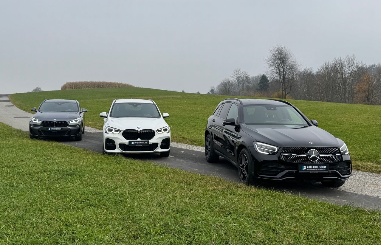 Drei Autos sind auf einer Straße geparkt: ein weißer BMW X1, ein schwarzer Mercedes-Benz GLC und ein graues Auto. Sie stehen auf einer Straße mit Rasen auf beiden Seiten.