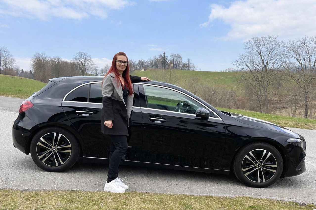 A woman with red hair and glasses is standing next to a black car on a road.