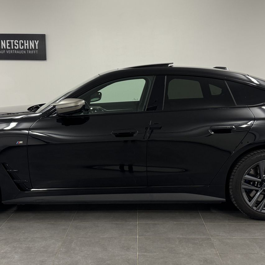 A black BMW car is parked in a showroom. A sign reading 'NETSCHNY AUF VERTAUEN TRIFFT' is on the wall.