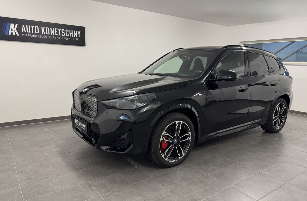A black BMW X1 SUV is parked inside a showroom with a white wall and a sign that reads 'Netschny'. The car has a sleek design and a shiny finish.