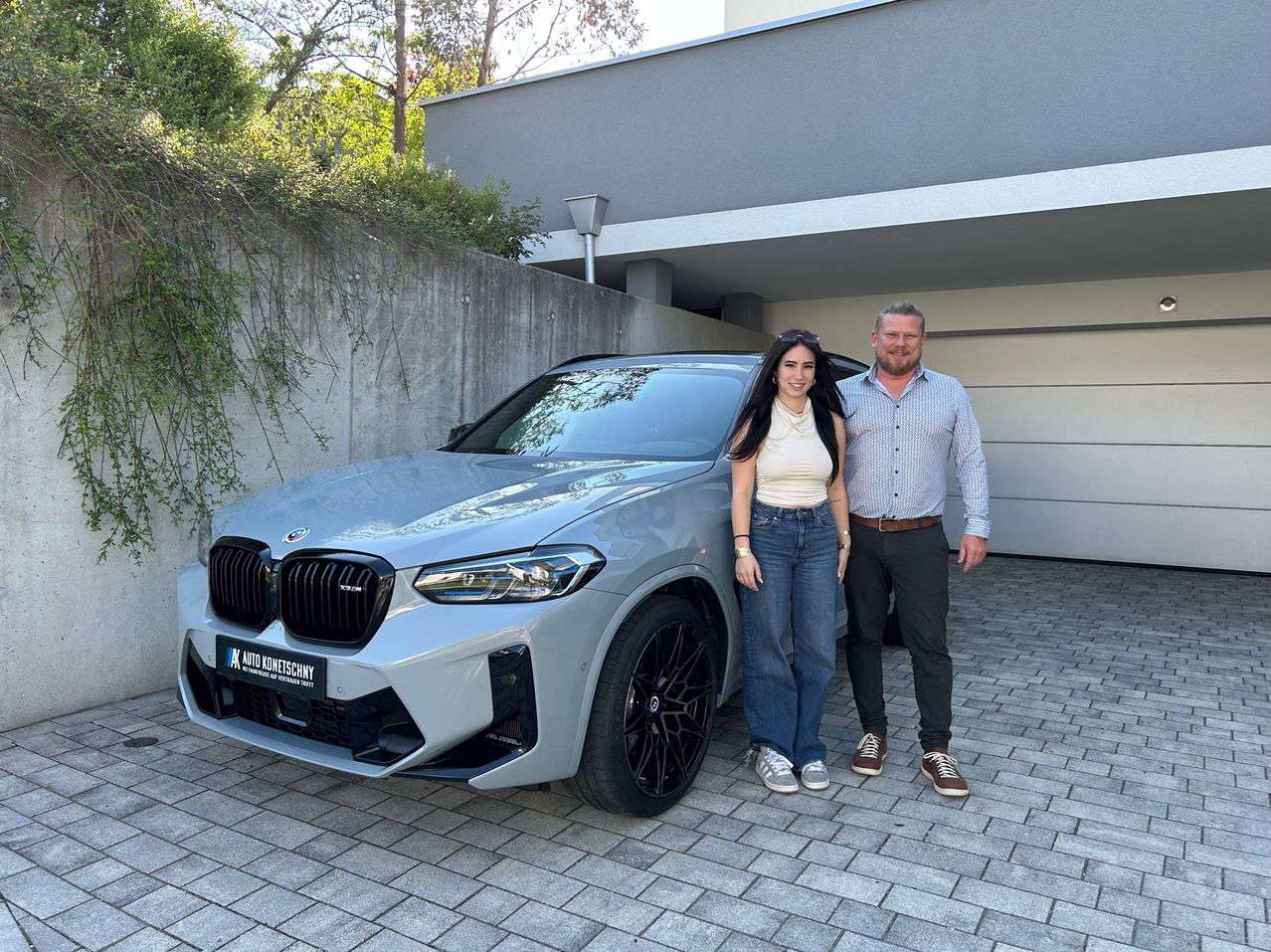 Ein Mann und eine Frau stehen vor einem silbernen BMW SUV mit Nummernschild auf einer gepflasterten Einfahrt an einem sonnigen Außenstandort.