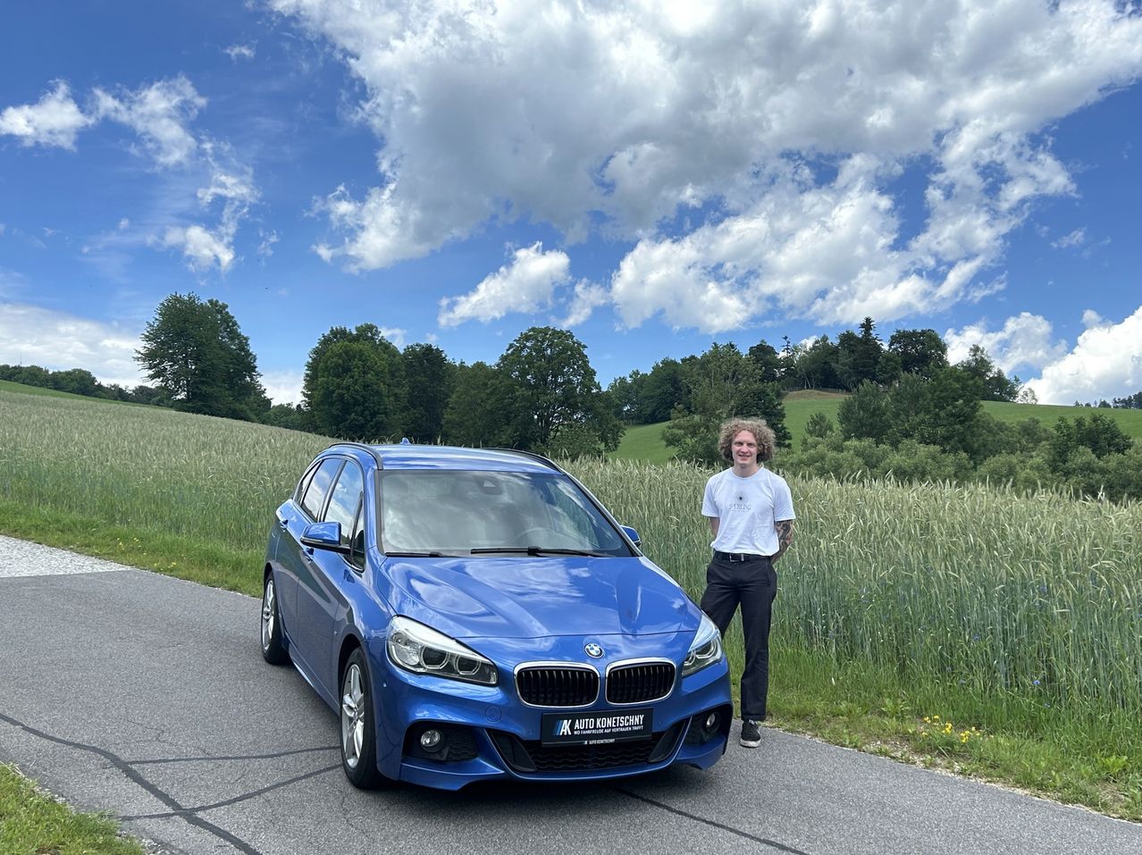 Eine Person steht neben einem blauen BMW auf einer Landstraße unter einem teilweise bewölkten Himmel.
