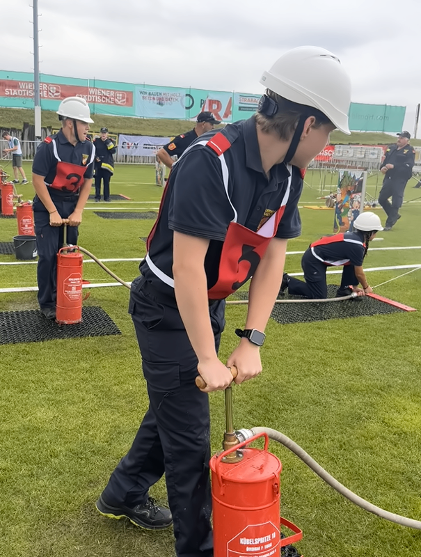 Eine Gruppe von Feuerwehrleuten in Schutzkleidung übt Feuerlöschtechniken auf einem Feld. Eine Person verwendet einen Feuerlöscher, während andere beobachten.