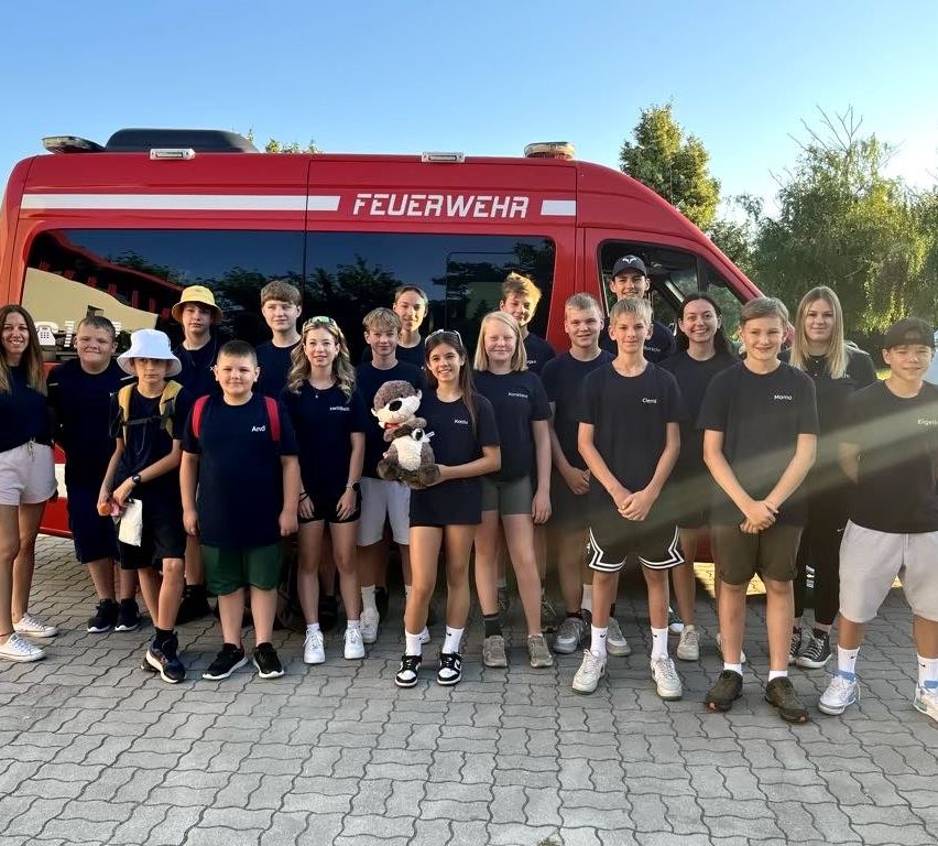 Eine Gruppe von Kindern und Jugendlichen steht vor einem roten Feuerwehrauto. Sie tragen alle passende T-Shirts, und einer hält ein Kuscheltier. Bäume und ein klarer Himmel sind im Hintergrund.