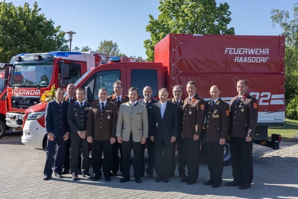 A group of men in uniforms stand in front of fire trucks. Some wear medals. The trucks are red.