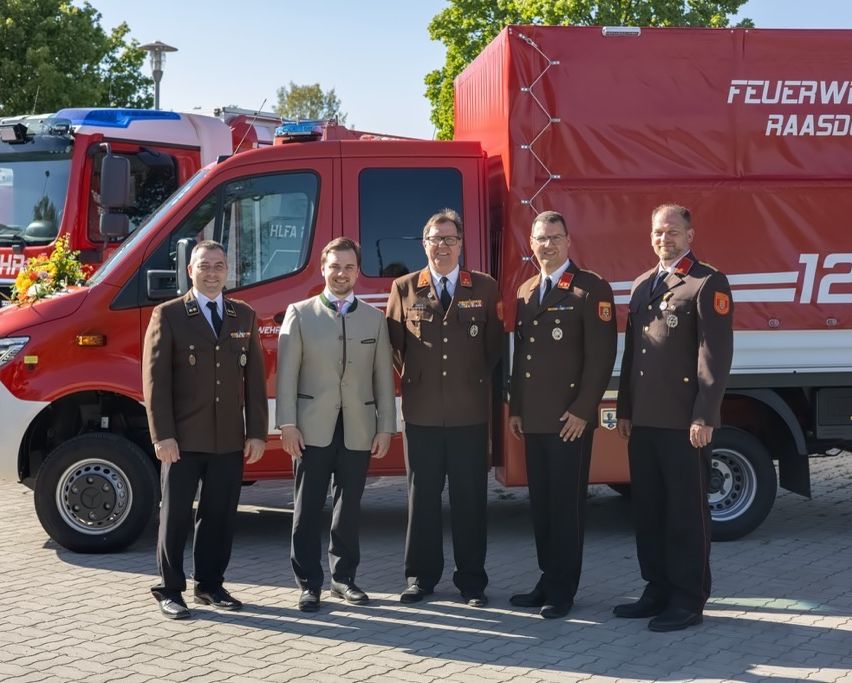 Five men in uniforms, including military and firefighter attire, stand in front of a red fire truck with 'FEUER' written on it.