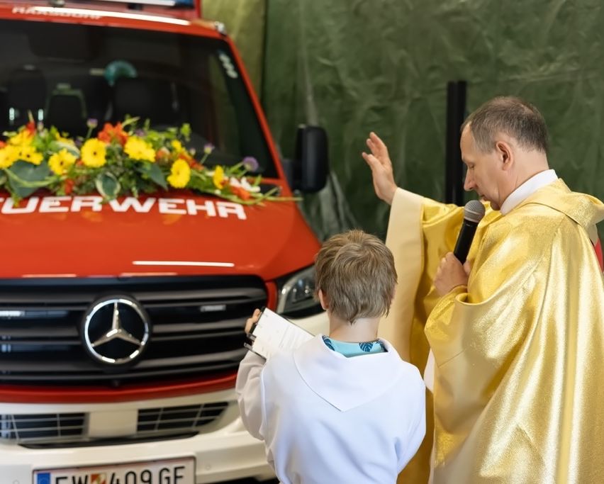 A priest blesses a fire truck with flowers. A boy in white stands nearby holding a book.