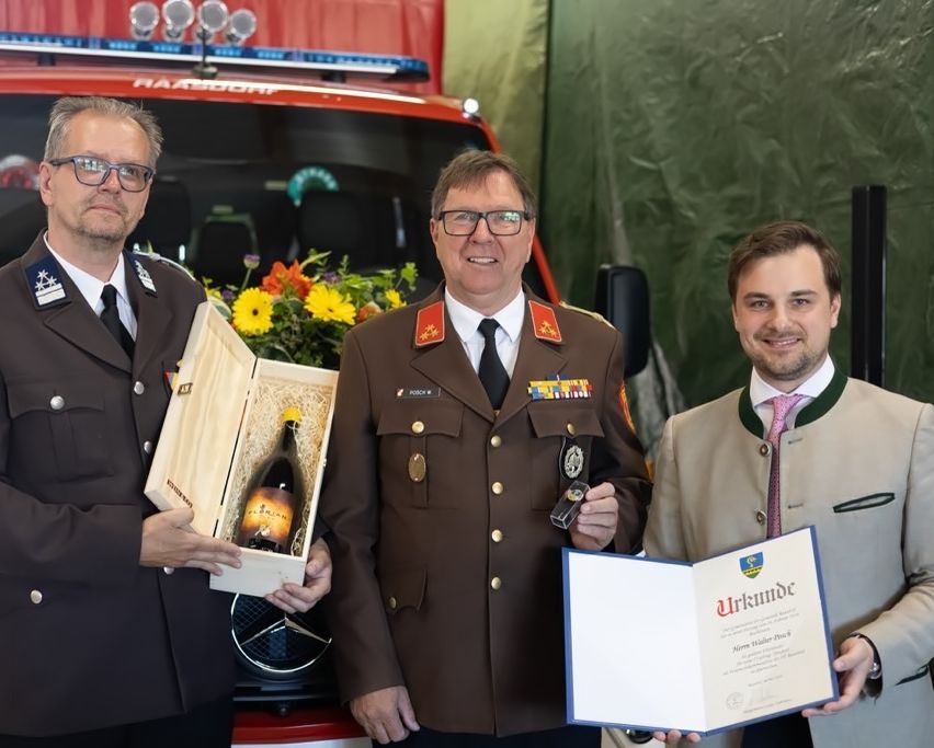 Three men stand in front of a red fire truck. One holds a bottle, another holds a certificate, and the third holds a bouquet of flowers. They all wear uniforms.