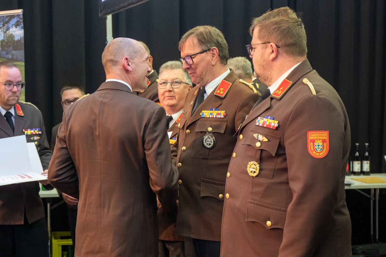 Three military officers in brown uniforms with medals and stars are conversing. One officer has a beard, and the others wear glasses. There are people behind them.