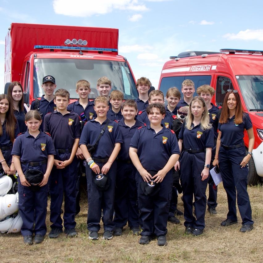 A group of young people dressed in navy uniforms with badges, standing in front of two fire trucks on a sunny day.