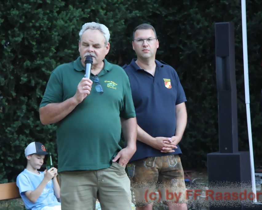 Two men stand outdoors; one speaks into a microphone, while another stands behind him. A boy sits on the ground nearby, holding a bottle. Behind them is a speaker.