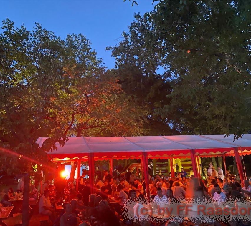 A large crowd of people are gathered under a red tent with a stage. They are sitting at tables and standing, enjoying a social event. Trees surround the area.