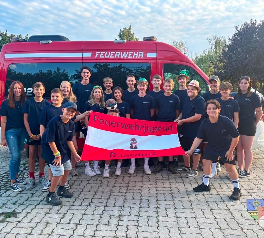 Eine Gruppe von Kindern und Jugendlichen in passenden blauen T-Shirts posiert für ein Foto vor einem roten Feuerwehrauto mit den Worten 'Feuerwehr' darauf. Sie halten eine rote Fahne mit einem weißen Logo und den Worten 'Feuerwehrjugend'.