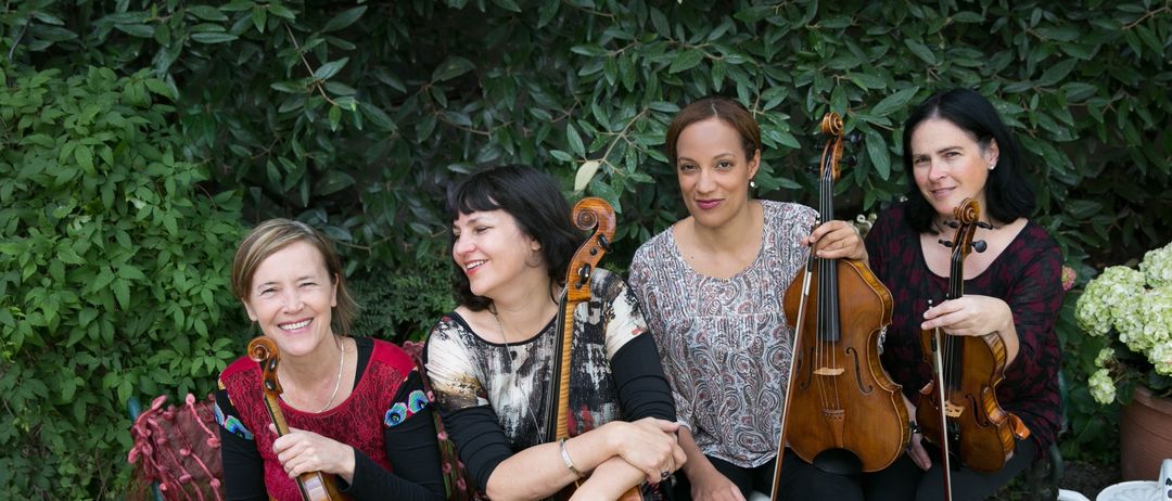 Vier Musikerinnen, jede mit einem Streichinstrument, sitzen in einer Außenumgebung. Zwei von ihnen lächeln, während eine von ihnen einen neutralen Ausdruck hat. Sie sind von üppiger Grünfläche und einem dekorativen Tisch mit einer Schüssel umgeben.