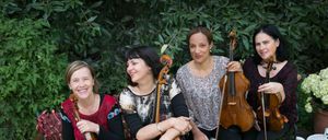 Vier Musikerinnen, jede mit einem Streichinstrument, sitzen in einer Außenumgebung. Zwei von ihnen lächeln, während eine von ihnen einen neutralen Ausdruck hat. Sie sind von üppiger Grünfläche und einem dekorativen Tisch mit einer Schüssel umgeben.