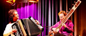 Two musicians perform on stage under spotlights. One plays a sitar while the other plays an accordion. They sit next to each other, focused on their instruments.