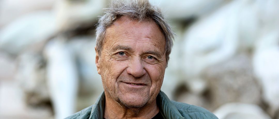 Ein älterer Mann mit grauem Haar lächelt, trägt ein schwarzes T-Shirt und eine grüne Jacke. Der Hintergrund ist unscharf, was auf einen sonnigen Außenbereich hindeutet.