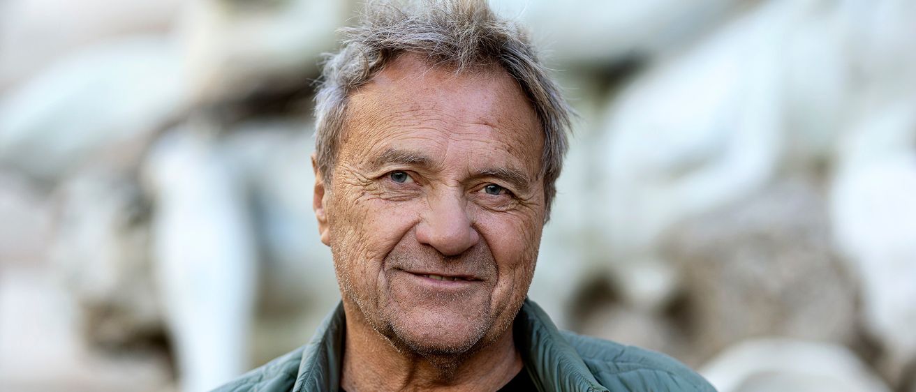 Ein älterer Mann mit grauem Haar lächelt, trägt ein schwarzes T-Shirt und eine grüne Jacke. Der Hintergrund ist unscharf, was auf einen sonnigen Außenbereich hindeutet.