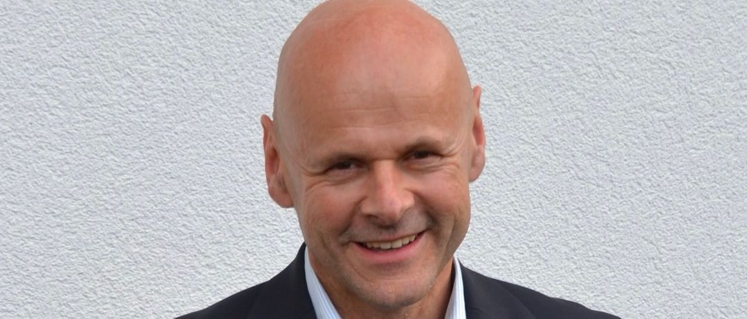 Ein glatzköpfiger Mann lächelt und trägt einen Anzug mit einem blauen Hemd darunter. Er steht vor einer hellen Wand.