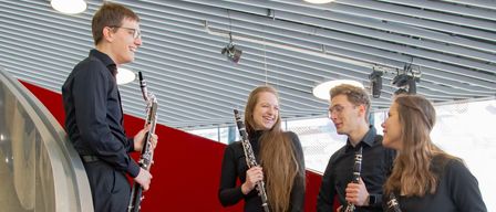 Vier Musiker, in Schwarz gekleidet, stehen auf roten Stufen und lächeln. Jeder hält ein anderes Holzblasinstrument. Die Umgebung ist modern und architektonisch.