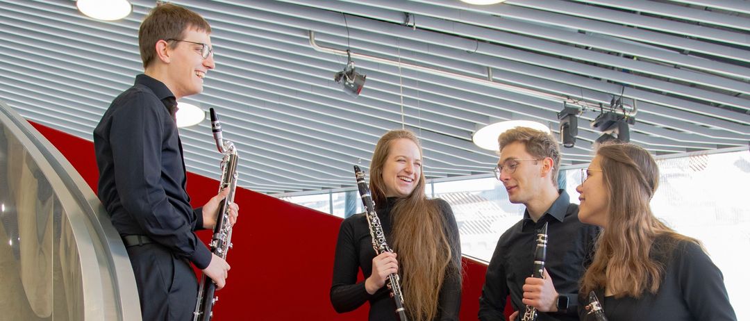 Vier Musiker, in Schwarz gekleidet, stehen auf roten Stufen und lächeln. Jeder hält ein anderes Holzblasinstrument. Die Umgebung ist modern und architektonisch.