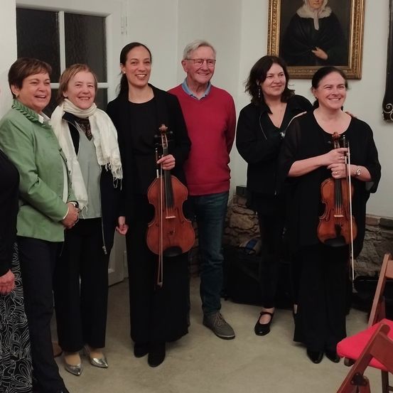 Eine Gruppe von Menschen, darunter zwei Frauen mit Violinen, lächelt für ein Foto in einem Raum. Der Mann in der Mitte trägt eine Brille und ein rotes Hemd. Hinter ihnen ist ein Bilderrahmen an der Wand angebracht.