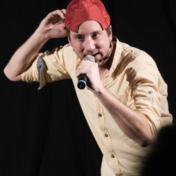 Ein Mann in einer roten Kappe und einem gelben Hemd spricht in ein Mikrofon auf der Bühne.